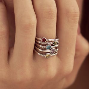 Puede incluir: Un conjunto de cinco anillos de plata, cada uno con una pequeña piedra preciosa de diversas formas y colores, incluyendo rojo, verde, azul, morado y amarillo, con diminutas piedras transparentes sobre finas bandas de plata pulida.