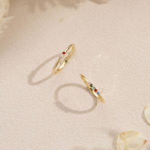 Peut inclure: Deux bagues fines en or, l'une ornée de trois petites pierres précieuses multicolores et l'autre de deux petites pierres précieuses multicolores. Les bagues sont posées sur une surface texturée avec le texte WestMonicaArts en bas au centre.
