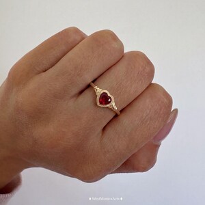 Peut inclure: Une bague en or avec une pierre précieuse rouge en forme de cœur au centre. Le sertissage présente un bord perlé décoratif autour de la pierre et un anneau fin en or poli. Le texte WestMonicaArts est visible au bas de l'image.