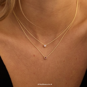 May include: A layered gold necklace set featuring two delicate chains. The shorter chain holds a small, light pink bezel-set gemstone, while the longer chain features a small, deep red bezel-set gemstone. Both pendants are circular in shape.