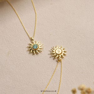 Könnte beinhalten: Zwei goldene Halsketten mit sonnenförmigen Anhängern. Ein Anhänger enthält in der Mitte einen runden, hellblauen Edelstein, während der andere Anhänger den eingravierten Buchstaben S in der Mitte trägt. Beide Anhänger hängen an dünnen Goldketten.