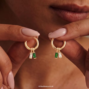 May include: A pair of gold hoop earrings, each featuring two dangling baguette-cut charms. One charm is clear and the other is emerald green. The earrings are held up, displaying the circular gold hoops and the small, rectangular gemstone pendants.