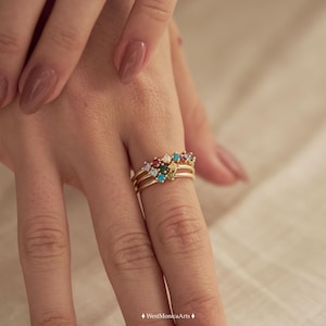 May include: A gold ring featuring three thin bands joined together, topped with a cluster of small, multi-colored gemstones including red, blue, green, yellow, and purple stones arranged in a triangular pattern on the finger.