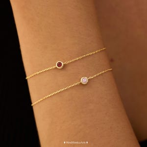 May include: Two delicate gold chain bracelets, each featuring a single bezel-set gemstone. The top bracelet displays a small, round, deep red stone, while the bottom bracelet features a small, round, light pink stone, both set in gold metal.
