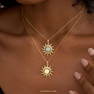 Peut inclure: Deux colliers dorés avec des pendentifs en forme de soleil. Le pendentif supérieur contient une petite pierre ronde bleu clair en son centre. Le pendentif inférieur présente un centre circulaire gravé de la lettre majuscule S.