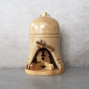 May include: A wooden Christmas bauble shaped like a bell with a nativity scene inside. The bauble is painted in shades of brown and features a star, a manger, and figures of Mary, Joseph, and a sheep.