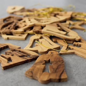 May include: A pile of wooden Christmas decorations in various shapes, including bells, stars, and trees. The decorations are made of light brown wood with a natural grain pattern. They are strung together with gold string.