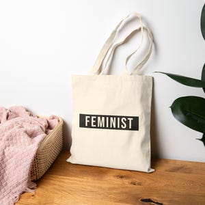 Könnte beinhalten: Eine cremefarbene Stofftasche aus Segeltuch mit zwei langen Schulterriemen und einem rechteckigen schwarzen Grafikdruck in der Mitte. Das Wort FEMINIST ist in fetten, weißen, serifenlosen Großbuchstaben in dem schwarzen Block gedruckt.