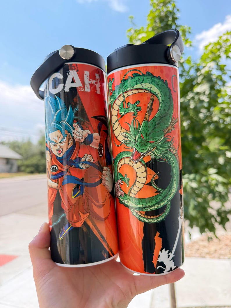 May include: Two cylindrical metal water bottles with black screw-top lids. The left bottle features a blue-haired character in an orange gi with the text CAH. The right bottle displays a large green dragon coiled against an orange background.