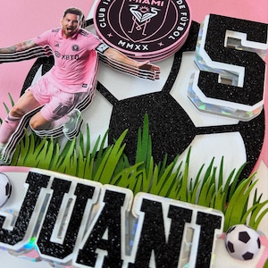 May include: A pink and black soccer-themed cake topper featuring a soccer player in a pink uniform. The topper includes the text "JUANI" and a soccer ball, with a "5" and a team logo.