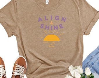 May include: A tan t-shirt with the words "ALIGN & SHINE" in purple and a yellow sun graphic. The shirt is styled with brown sneakers, sunglasses, blue jeans, and white roses.
