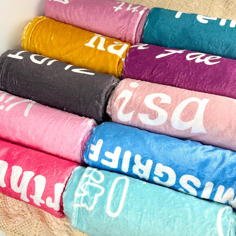 Puede incluir: Una colecci&oacute;n de mantas enrolladas en varios colores, incluyendo rosa, amarillo y azul. Cada manta muestra un dise&ntilde;o de texto blanco. Las mantas est&aacute;n apiladas, mostrando una variedad de tonos y patrones.