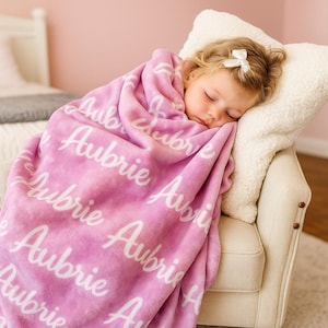 Puede incluir: Un ni&ntilde;o dormido envuelto en una suave manta rosa. La manta est&aacute; personalizada con el nombre "Aubrie" en una escritura blanca repetida. El ni&ntilde;o descansa sobre una silla color crema con una almohada blanca esponjosa.