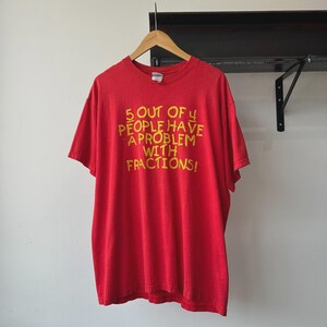 May include: A bright red short-sleeved t-shirt featuring yellow screen-printed text that reads 5 OUT OF 4 PEOPLE HAVE A PROBLEM WITH FRACTIONS! The shirt is displayed on a wooden hanger, showcasing a crew neckline and a standard straight hem.