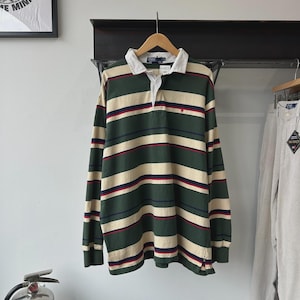 May include: A long-sleeved rugby shirt with horizontal stripes in green, beige, and red. The shirt has a white collar and a small red logo on the chest. The shirt is hanging on a wooden hanger.