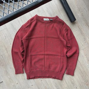 May include: A red sweater with a grid pattern. The sweater has a crew neck and long sleeves. The fabric appears to be a knit material. The sweater is lying on a light-colored wooden surface.