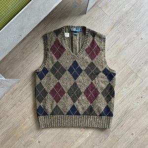 May include: A sleeveless V-neck sweater vest featuring a brown, burgundy, and navy blue argyle diamond pattern. The knit garment has a ribbed hem and neckline, with a Polo by Ralph Lauren label at the inner collar and a size L tag attached.