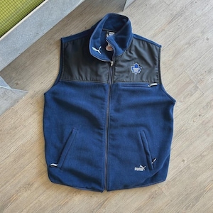 May include: A navy blue fleece vest featuring a black panel across the upper chest and shoulders. The garment includes a full-length front zipper, two side pockets, and a small chest pocket. A Toronto Maple Leafs logo and a white Puma logo are visible.