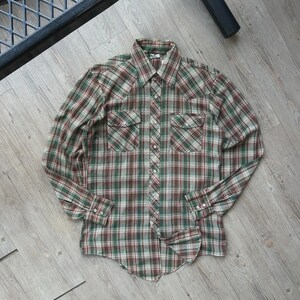 May include: A long-sleeved plaid shirt in shades of green, brown, and white. The shirt has two chest pockets, a button-down front, and buttoned cuffs. The shirt is lying flat on a wooden surface.