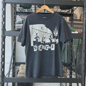 May include: Dark gray t-shirt with a graphic print featuring the band Static-X. The design includes band members, text, and a logo. The shirt is hanging on a wooden hanger.