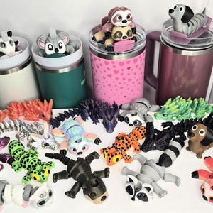 May include: A collection of animal-themed tumbler toppers and figurines. The toppers include a possum, a mouse, a sloth, and a dachshund. The figurines include dragons, raccoons, and other creatures. The tumblers are white, green, pink, and purple.
