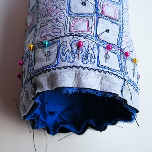 May include: A gray fabric tube with blue embroidery and a blue lining. The fabric is pinned together with colorful pins.