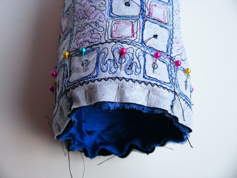 May include: A gray fabric tube with blue embroidery and a blue lining. The fabric is pinned together with colorful pins.