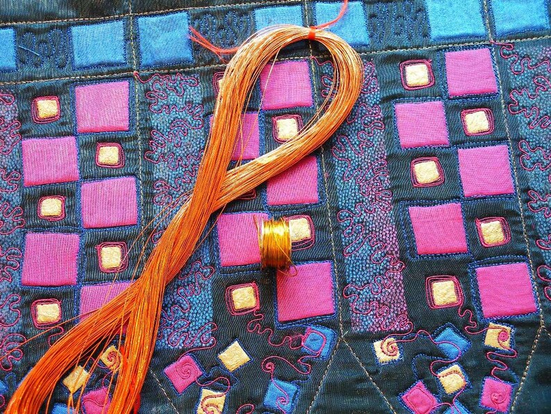 May include: A close-up of a copper wire coil on a fabric background with a geometric pattern of pink and blue squares. The fabric has a textured surface and is stitched with a decorative design.