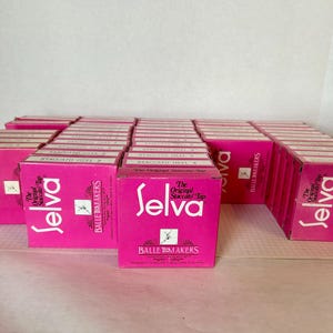 May include: A collection of bright pink cardboard boxes for Selva The Original Staccato Tap ballet makers. Each box features white text with the brand name Selva, the product description, and a small graphic of a dancer in a pose on the front.