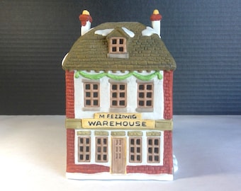 Può includere: Una miniatura in ceramica di un edificio con facciata in mattoni, tetto marrone e dettagli di neve. L'edificio ha più finestre e la scritta "M. Fezziwig Warehouse" su un cartello. Ghirlande verdi decorano la parte anteriore.