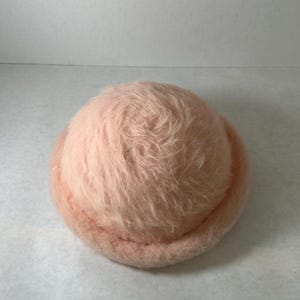 May include: A soft, fuzzy, pale pink hat with a rounded crown and a rolled brim. The texture of the hat is visibly shaggy and dense, featuring a uniform light peach-pink color throughout the entire surface of the rounded headwear piece.