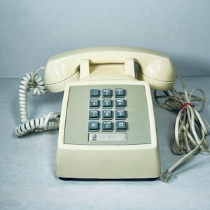 May include: A cream-colored landline telephone with a matching handset resting on top. The device features a gray keypad with twelve square buttons, including numbers, an asterisk, and a pound sign. An AT&T logo is visible below the keypad.