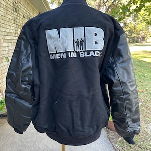 May include: Black varsity jacket with leather sleeves. The back of the jacket features the silver letters "MIB" above the words "MEN IN BLACK" and a silhouette of two figures.