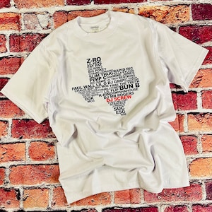 Può includere: T-shirt bianca con grafica nera e rossa a forma di Texas, piena di nomi. Il testo "DJ SCREW" è in rosso. La maglietta è realizzata in un materiale morbido.