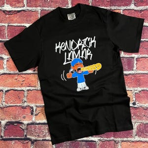 May include: Black t-shirt with the white text "Kendrick Lamar" and a cartoon illustration. The illustration depicts a person in a blue shirt and hat holding a yellow object with the word "MUSTARD".