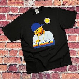 Può includere: T-shirt nero con grafica colorata di un uomo con berretto blu e catena d'oro. Il testo "DJ SCREW" è visualizzato in un font retrò. Un logo circolare è visibile in alto a destra.
