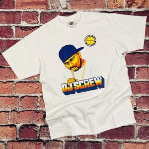 Può includere: T-shirt bianco con grafica di DJ Screw con berretto blu e catena d'oro. Il design include il testo "DJ SCREW" in un font stilizzato e un logo circolare con testo. La maglietta è esposta su uno sfondo di muro di mattoni.