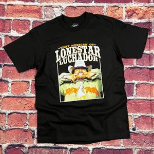 May include: Black t-shirt featuring a graphic of a man in a cowboy hat with the text "THAT MEXICAN OT LONESTAR LUCHADOR" in white and gold. The image is set against a brick wall background.
