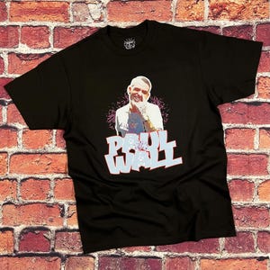Pode incluir: Camiseta preta com um gráfico de um homem e o texto "PAUL WALL" em uma fonte estilizada. O gráfico tem um esquema de cores rosa e azul. A camiseta é exibida em um fundo de parede de tijolos.