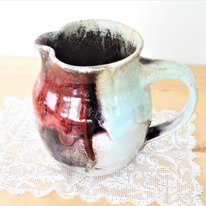 May include: A handmade ceramic pitcher with a unique glaze featuring shades of red, brown, and blue. The pitcher has a rounded body and a handle.