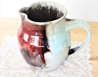 May include: A handmade ceramic pitcher with a unique glaze featuring shades of red, brown, and blue. The pitcher has a rounded body and a handle.