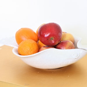 May include: A white ceramic fruit bowl with a wavy rim, filled with red apples and orange oranges.