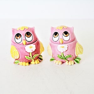May include: Two pink ceramic owl figurines with yellow accents and a white flower design. The owls have large eyes and are sitting on a white surface.