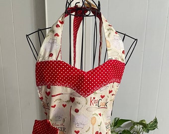 May include: A red and white polka dot apron with a halter neck strap. The apron has a cream background with a repeating pattern of cherries, hearts, and the text "Kiss the Cook" and "Bless the Baker".