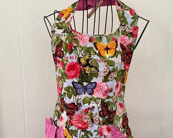 May include: A blue and pink floral apron with butterfly print and two pink pockets. The apron has a halter neck style and is made of cotton fabric.