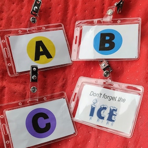 Puede incluir: Cuatro portatarjetas de plástico transparente con diferentes inserciones. Uno tiene un círculo amarillo con una "A" negra, otro un círculo azul con una "B" negra, y un tercero un círculo morado con una "C" negra. El cuarto tiene el texto "Don't forget the ICE".