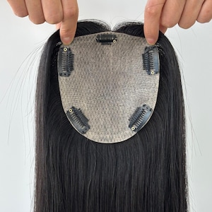 Puede incluir: Un postizo de cabello negro con una base de encaje y varios clips para sujetarlo. El cabello es largo y liso, con una textura natural. El postizo está diseñado para añadir volumen y cobertura.