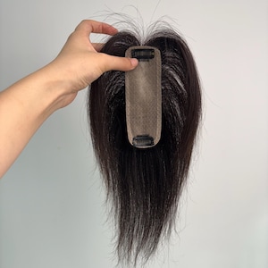 Pode incluir: Uma touca de cabelo castanho escuro com uma base de encaixe. A peruca é segurada contra um fundo branco. O cabelo é longo e liso, com um aspeto natural.