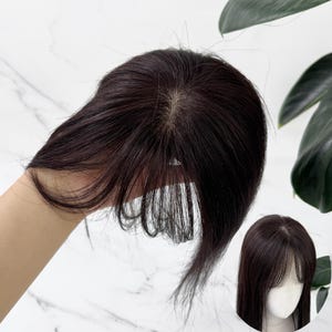 以下が含まれることがあります： 頭皮のような見た目のモノフィラメントベースと、薄く軽やかな前髪が特徴のダークブラウンのヘアトッパー。髪はストレートで滑らかであり、手で持った状態とマネキンの頭部に装着した状態の両方で示されている。
