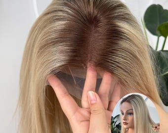 May include: A blonde wig with a dark brown root. The wig is held up by a hand, showing the lace front and the natural-looking hairline. The hair is straight and falls to the shoulders.